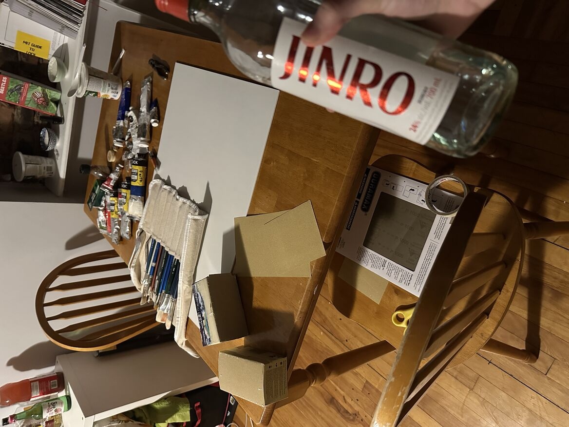 a messy dining table covering with paint brushes, sand paper, paint tubes, gesso and other materials. the photo is humorous with the photography holding a bottle of alcohol like a video game character holding a sword.