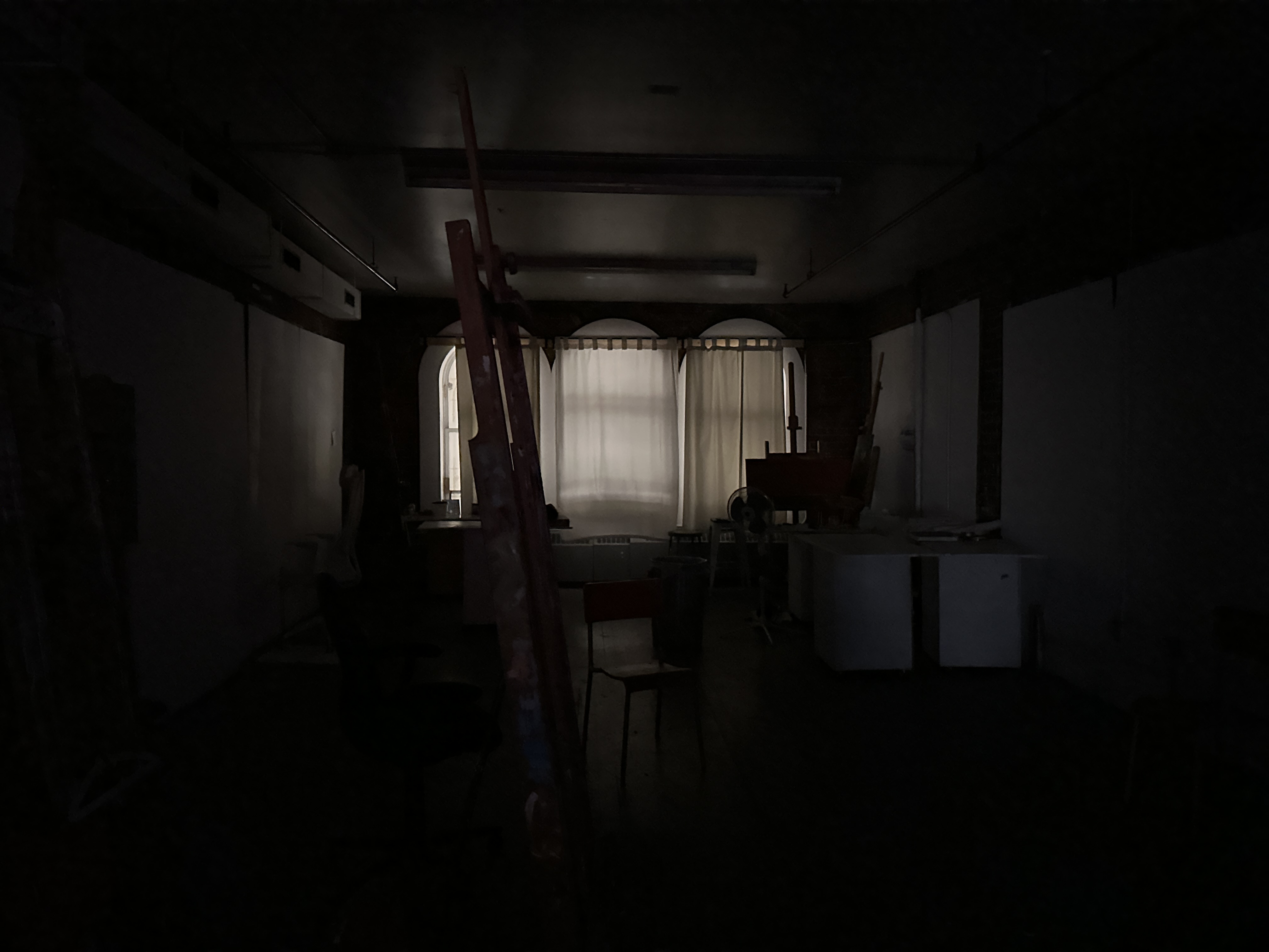 a darkly lit painting studio, large windows at the back with the curtains drawn. there are easels and paint boxes strawn across the room in the dark.